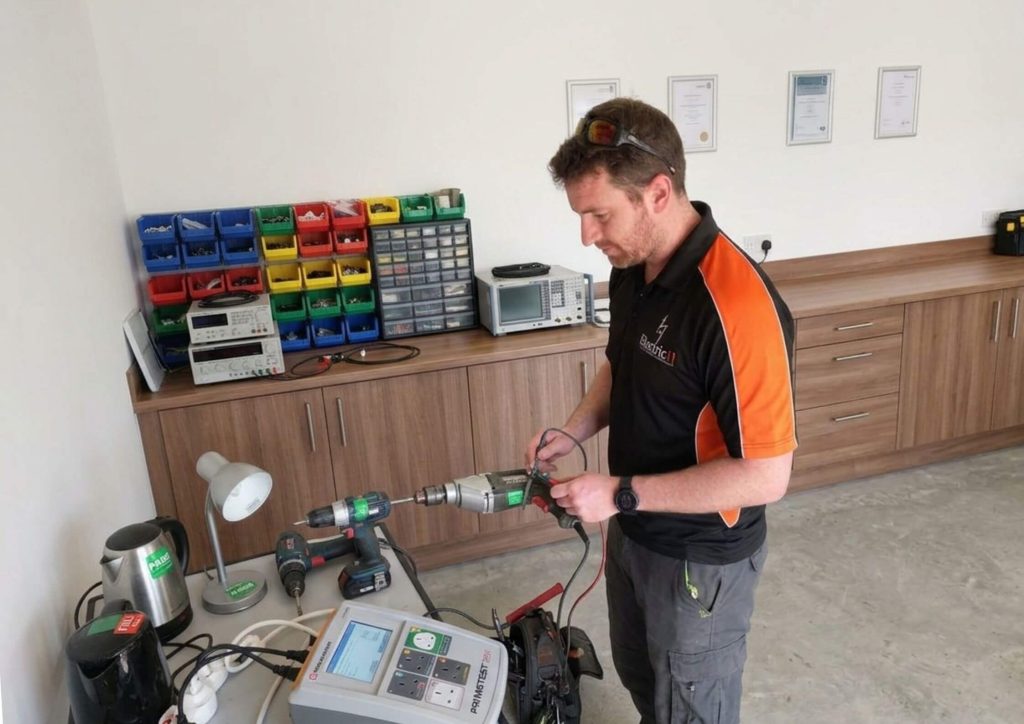 An electrician wearing a black and orange Electric 11 polo shirt stands at a wooden workbench in a bright, modern workshop. He is using a Seaward PrimeTest PAT tester to inspect a handheld power drill. The background features organized storage bins, electrical testing equipment, and framed certifications on a white wall.