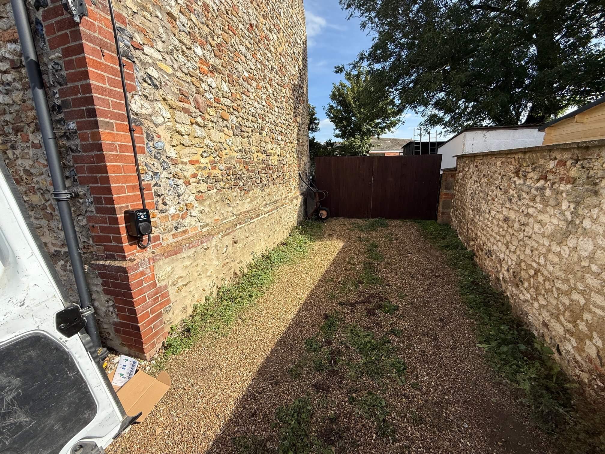 Gravel driveway between stone walls leading to a closed wooden gate, with a black Ohme electric vehicle charger mounted on the brick corner of the building on the left.