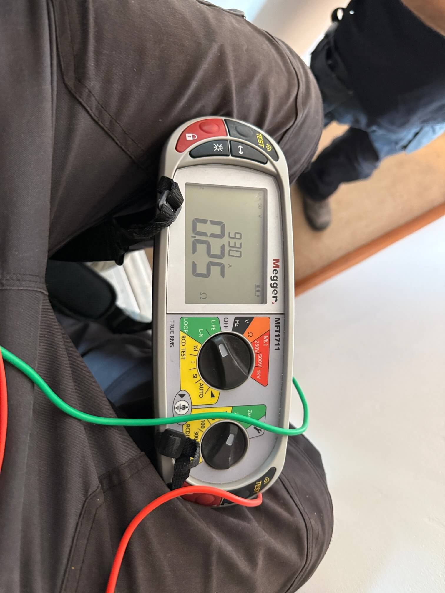 Megger multifunction electrical tester resting on a person’s lap, with a digital reading on its screen, two rotary controls, and red and green test leads attached.