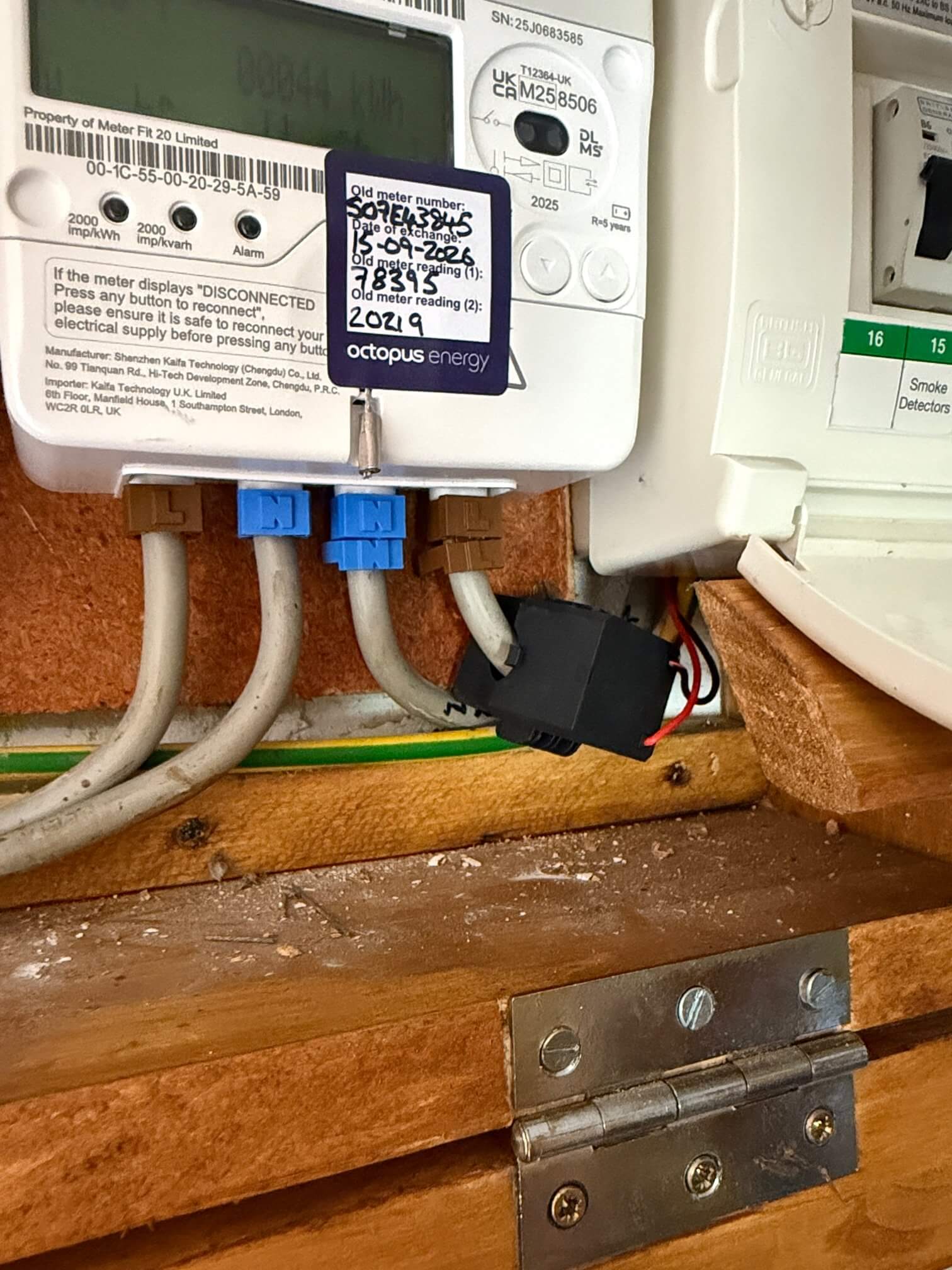 Close-up of cables beneath a digital electricity meter, with a black clamp sensor around one grey cable and thin red and black wires beside the consumer unit.