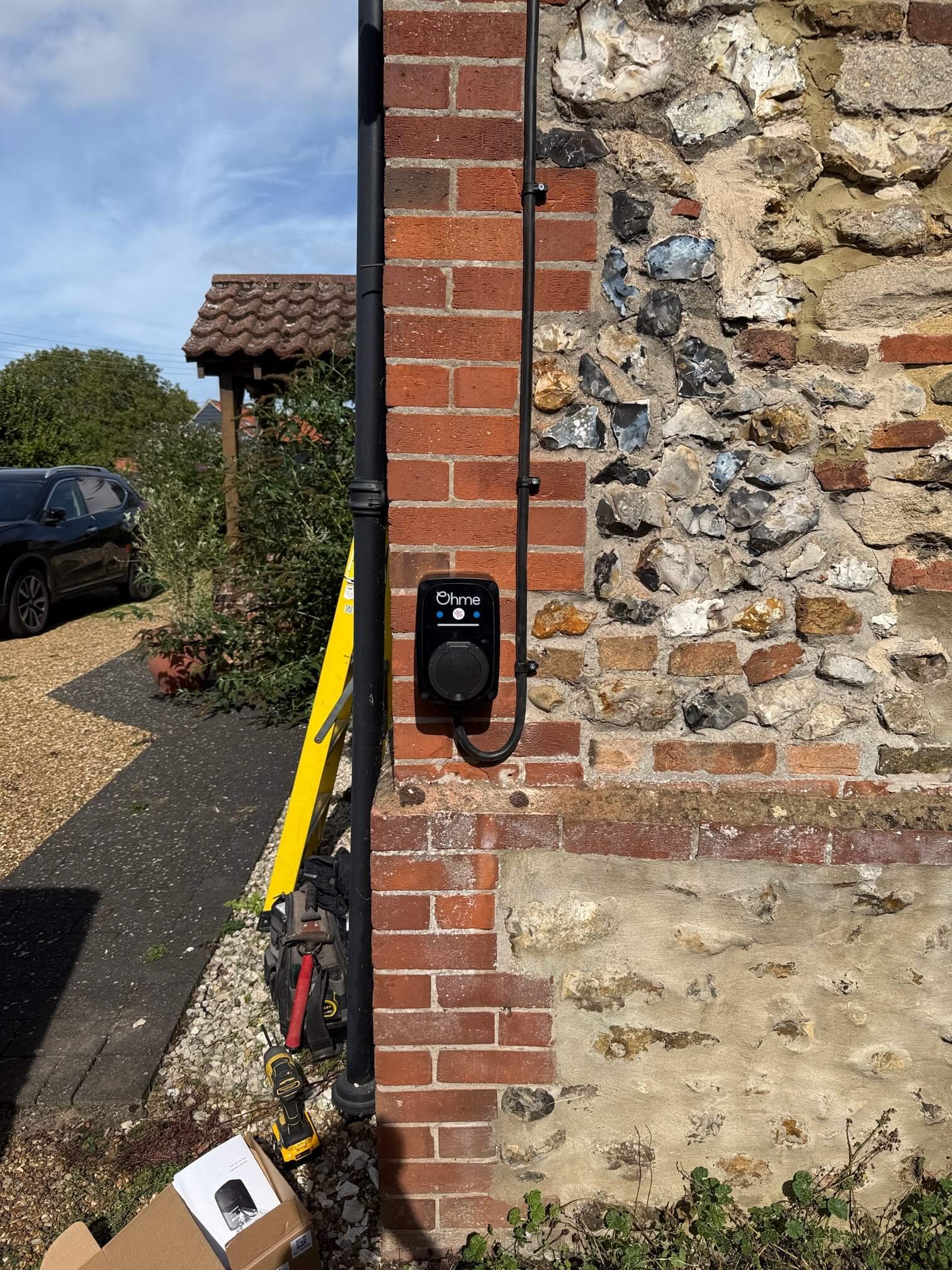 Black Ohme electric vehicle charger mounted on the brick edge of a stone exterior wall, with its cable running upward beside a drainpipe. Tools and packaging sit below.